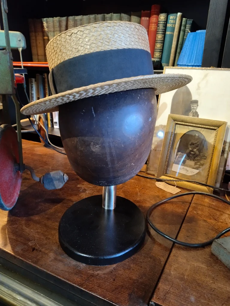 Das Foto zeigt einen Vintage-Hutständer aus Holz, der mit einem Strohhut versehen ist. Der Ständer steht auf einem hölzernen Ausstellungstisch. Die Szenerie strahlt eine nostalgische und klassische Atmosphäre aus, die den Charme und die Eleganz von Vintage-Dekor unterstreicht.