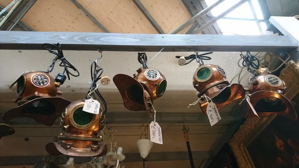 A collection of vintage diving helmets is displayed in an antique shop. These helmets are reminiscent of early 20th-century deep-sea diving gear, made from copper with green-tinted glass portholes and intricate metalwork. Each helmet hangs from the ceiling.