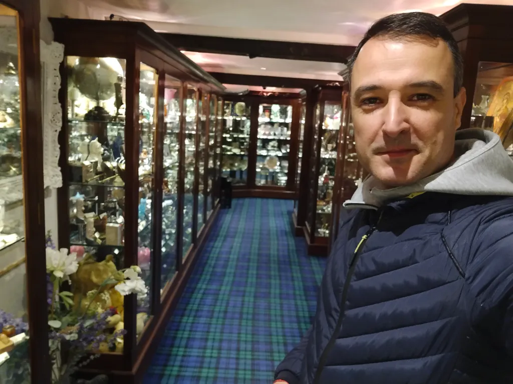 This is a photo of me in the showroom of Cromwell Antique Center. I am standing in a hallway lined with tall wooden cabinets filled with various antiques and collectibles. The floor is covered with a blue and green tartan-patterned carpet, adding to the elegant atmosphere of the showroom. The cabinets are well-lit, showcasing an array of items such as glassware, figurines, and other valuable objects. The overall ambiance is cozy and inviting, perfect for an antique hunting adventure.