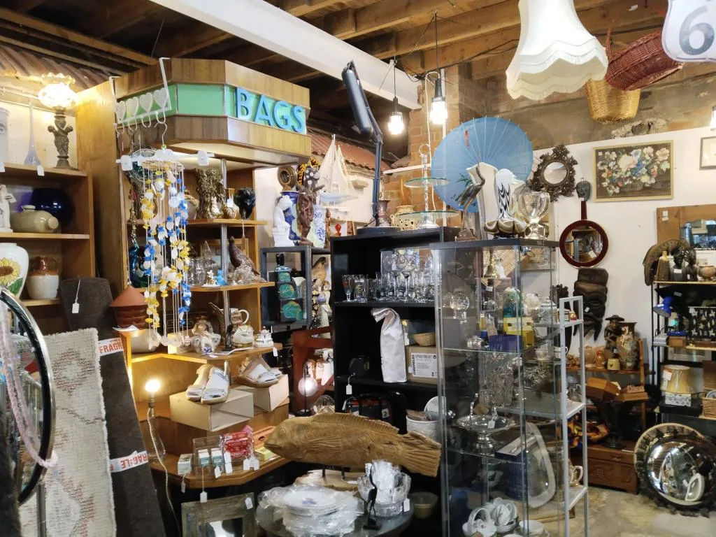 The interior of an antique shop is filled with a diverse array of vintage items. Shelves and display cases are packed with an eclectic mix of collectibles, including ceramic figurines, glassware, decorative plates, lamps, framed artwork, and various other curiosities.