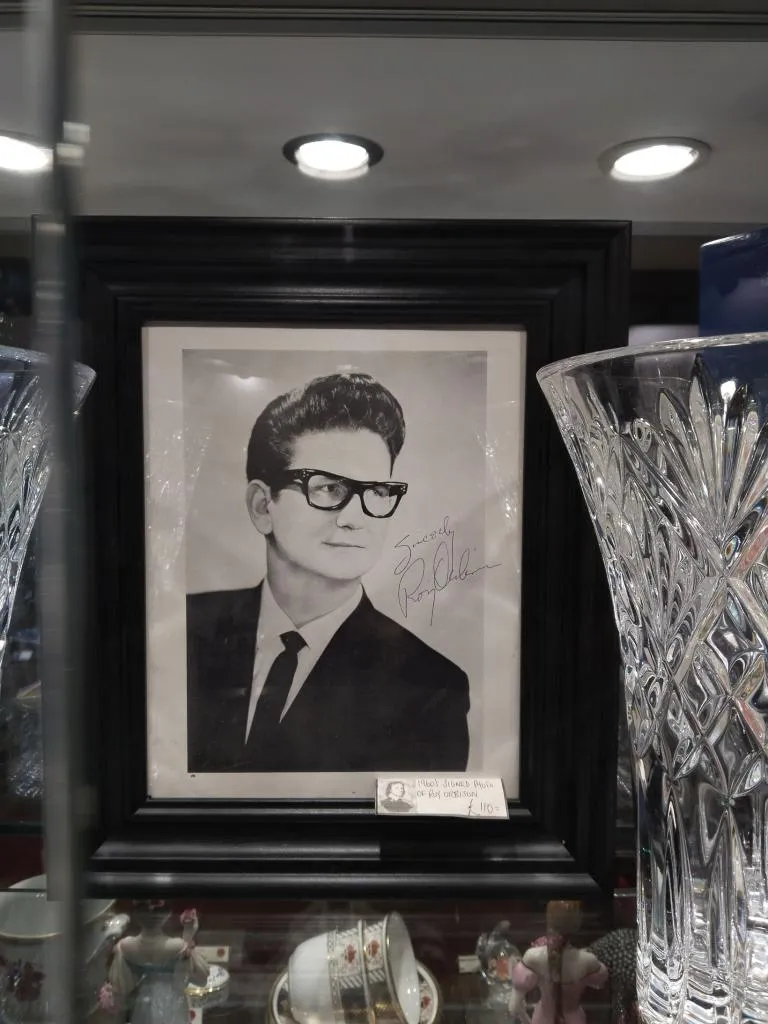 This is a signed photo of Roy Orbison. The black and white photograph is framed in a black frame and features Roy Orbison wearing his iconic glasses and a suit. His signature, 